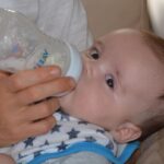 Un nourrisson allaite avec une biberne, soutenu par une main sur un coussin