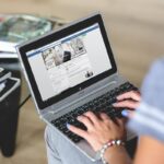 Une personne tape sur un clavier d'ordinateur portable à côté d'un carnet et d'une tasse de café blanche