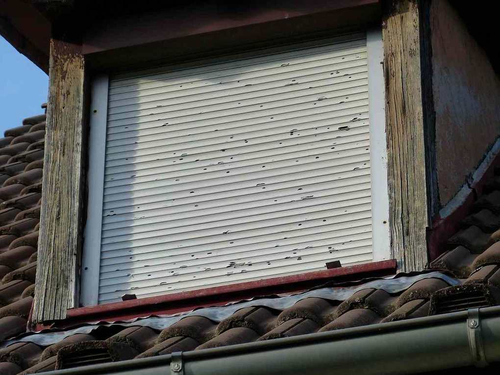 Volet roulant blanc fermé sur une fenêtre avec encadrement en bois usagé, toit en tuiles