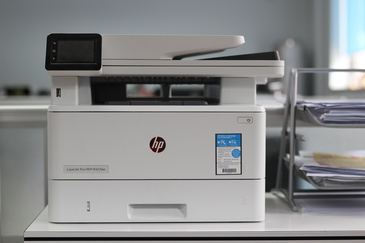 Imprimante multifonction HP blanche posée sur un meuble de bureau, photographiée de face Imprimante multifonction HP blanche posée sur un meuble de bureau, photographiée de face