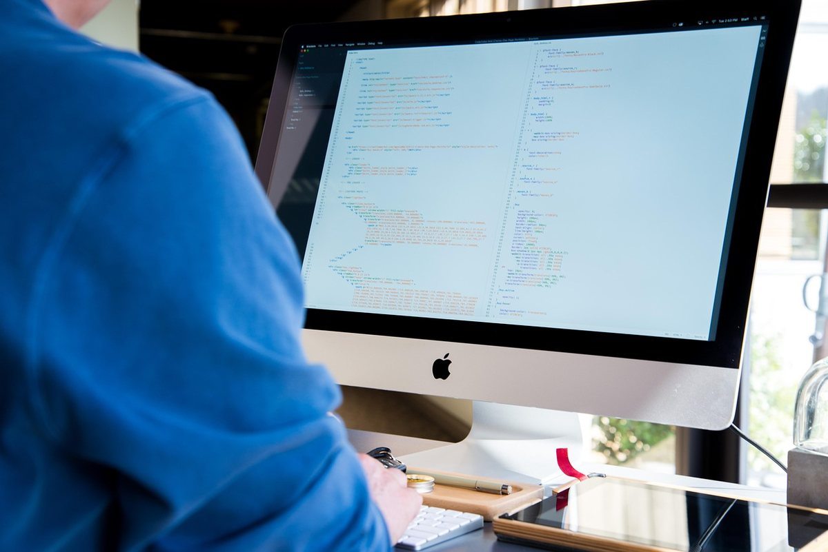 Individu en bleu devant un iMac affichant du texte, lumière ambiante Individu en bleu devant un iMac affichant du texte, lumière ambiante